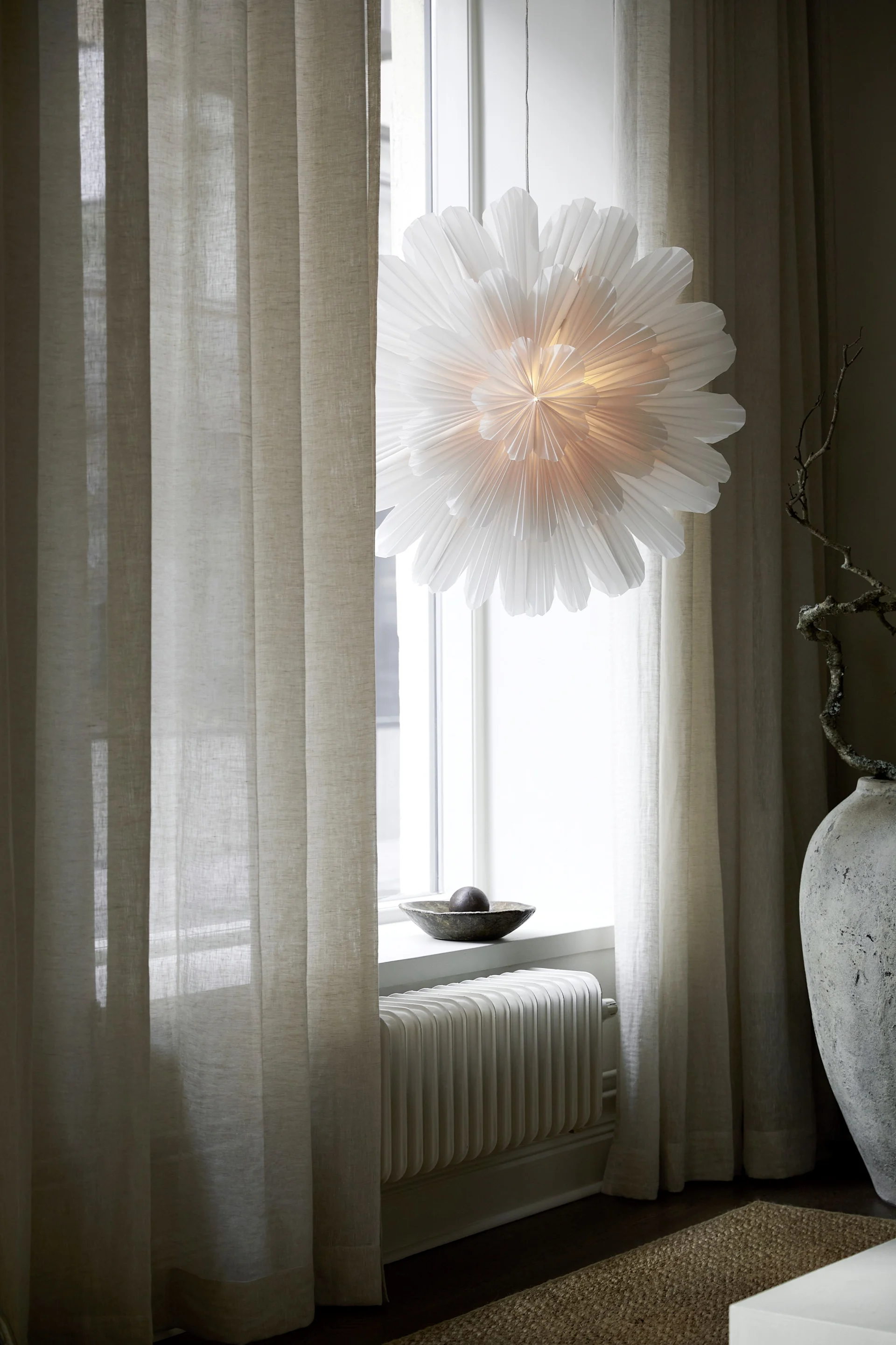 A white pleated pendant lamp hangs in a window, flanked by sheer curtains, above a radiator and bowl with a sphere.