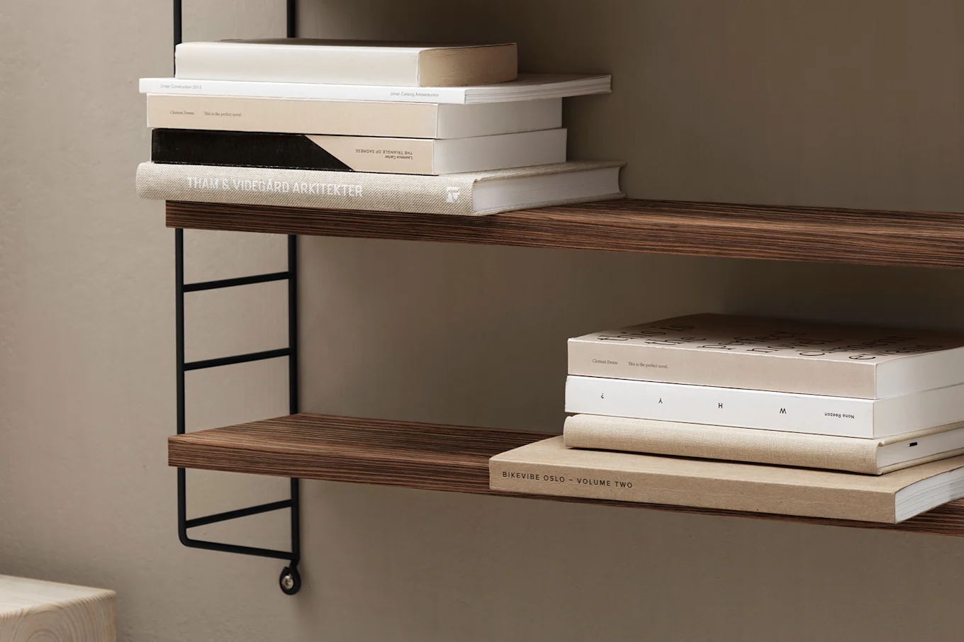 A wall-mounted shelving unit with two wooden shelves holding stacks of neutral-toned books against a beige wall.