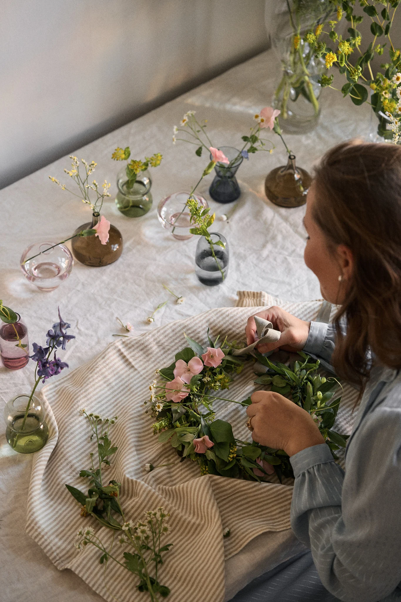 Add making a midsummer wreath to your summer bucket list ideas. Here you see a woman putting together a wreath surrounded by the mini Orrefors midsummer vases. 