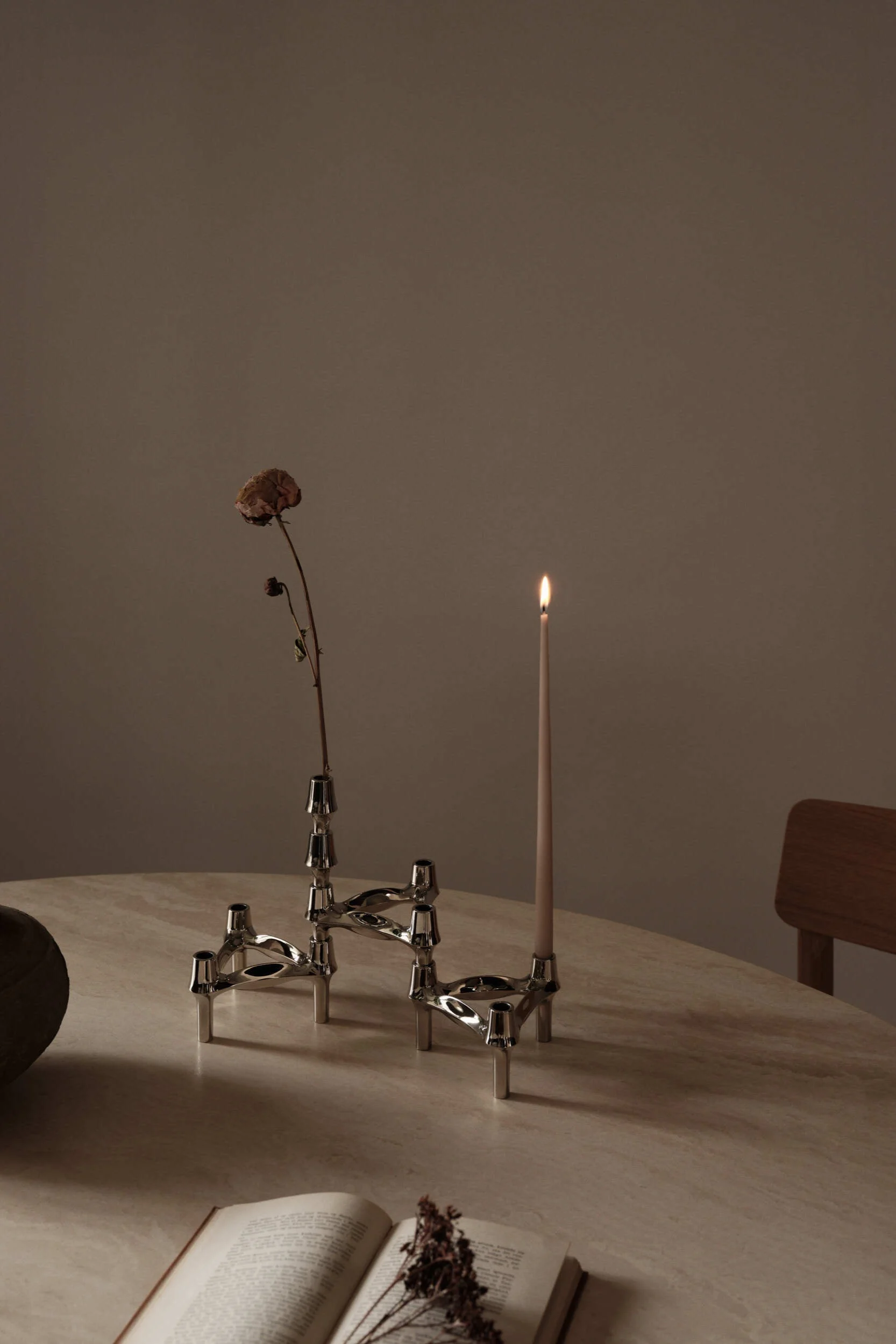 Two silver-toned modular candle holders on a round table, one with a lit candle, the other with a dried flower. An open book is visible in the foreground.