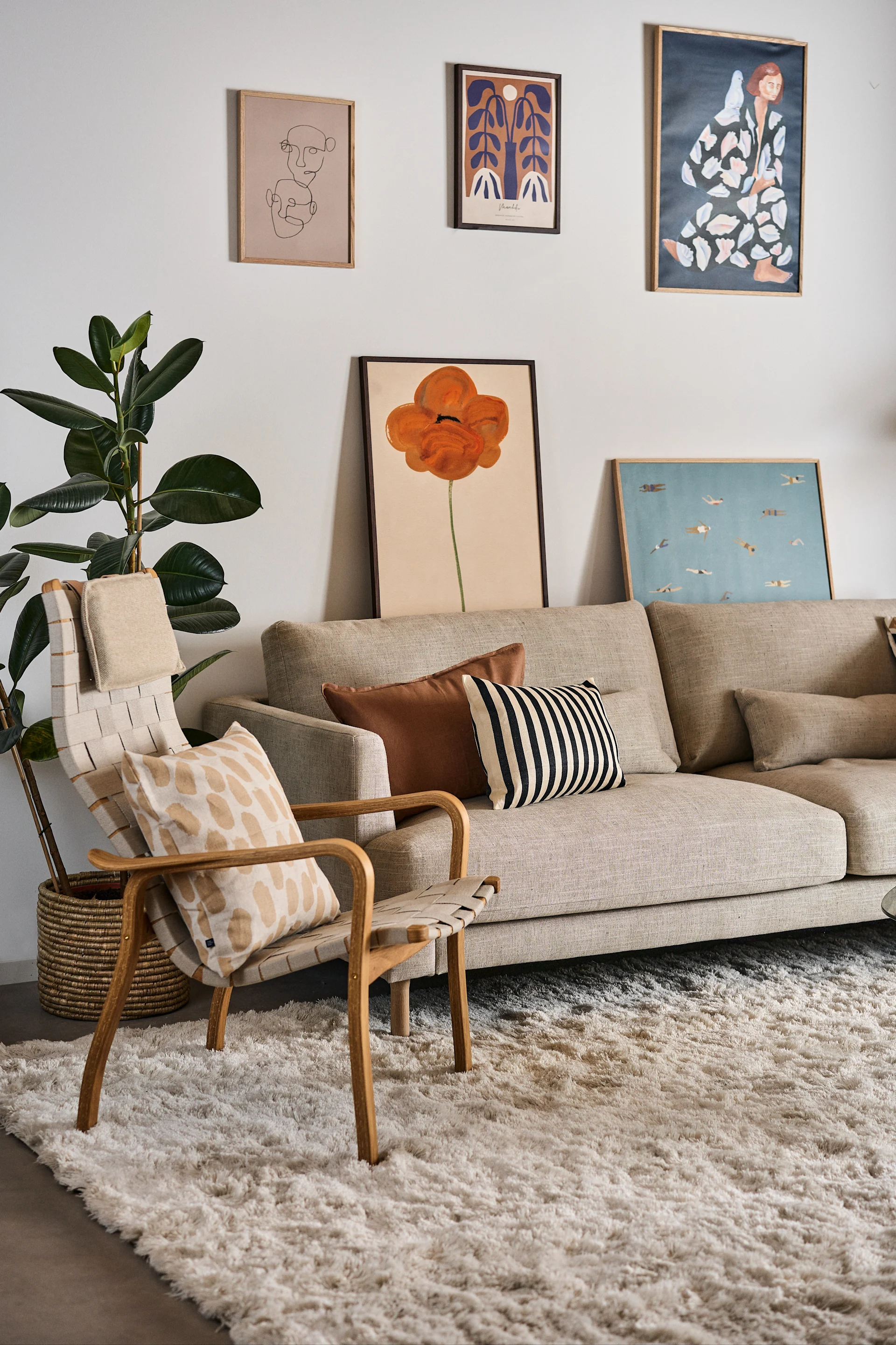 Modern living room with a beige sofa, woven armchair, large green plant, creamy rug, and several framed art prints.