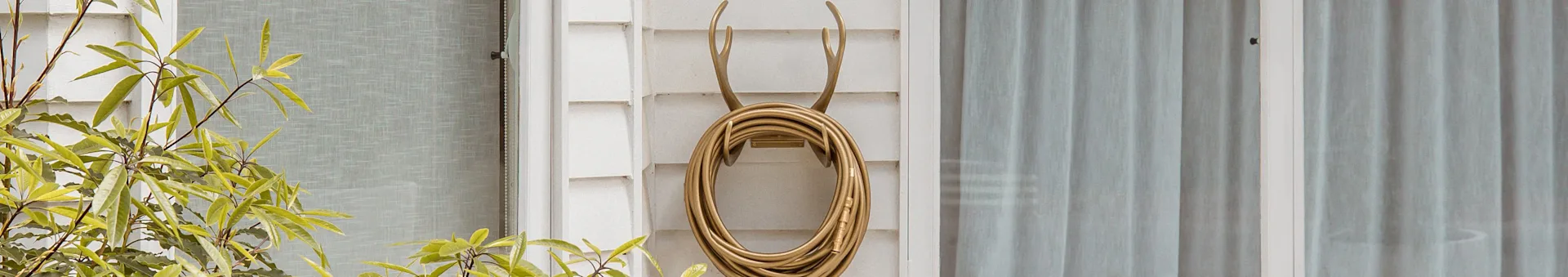 A gold coiled garden hose hangs on a decorative antler-shaped hook on a white wall, with green plants and a window nearby.