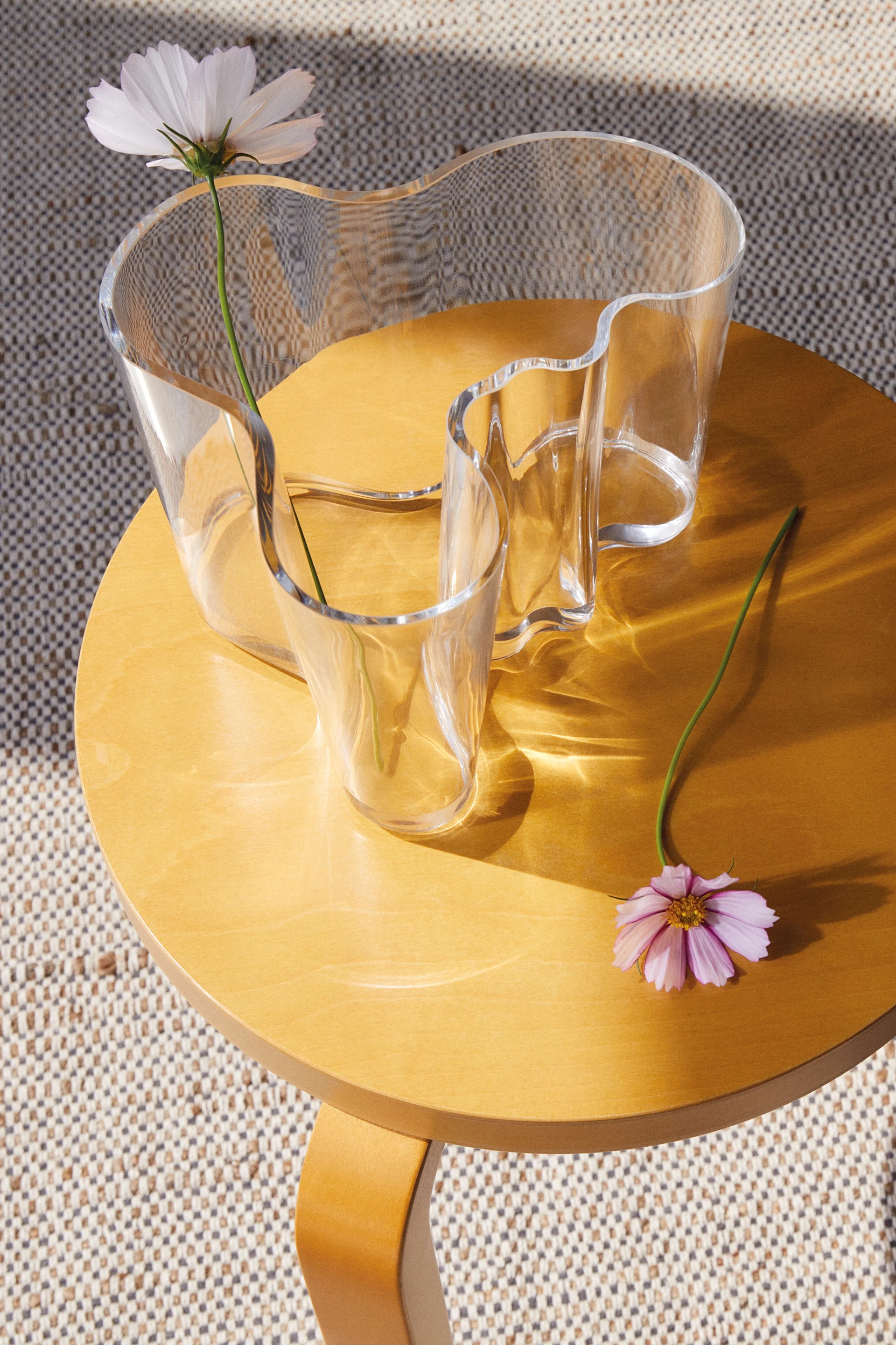 A clear, wavy glass vase holds a white flower on a round wooden table, with a pink flower lying beside it. Sunlight casts patterns.
