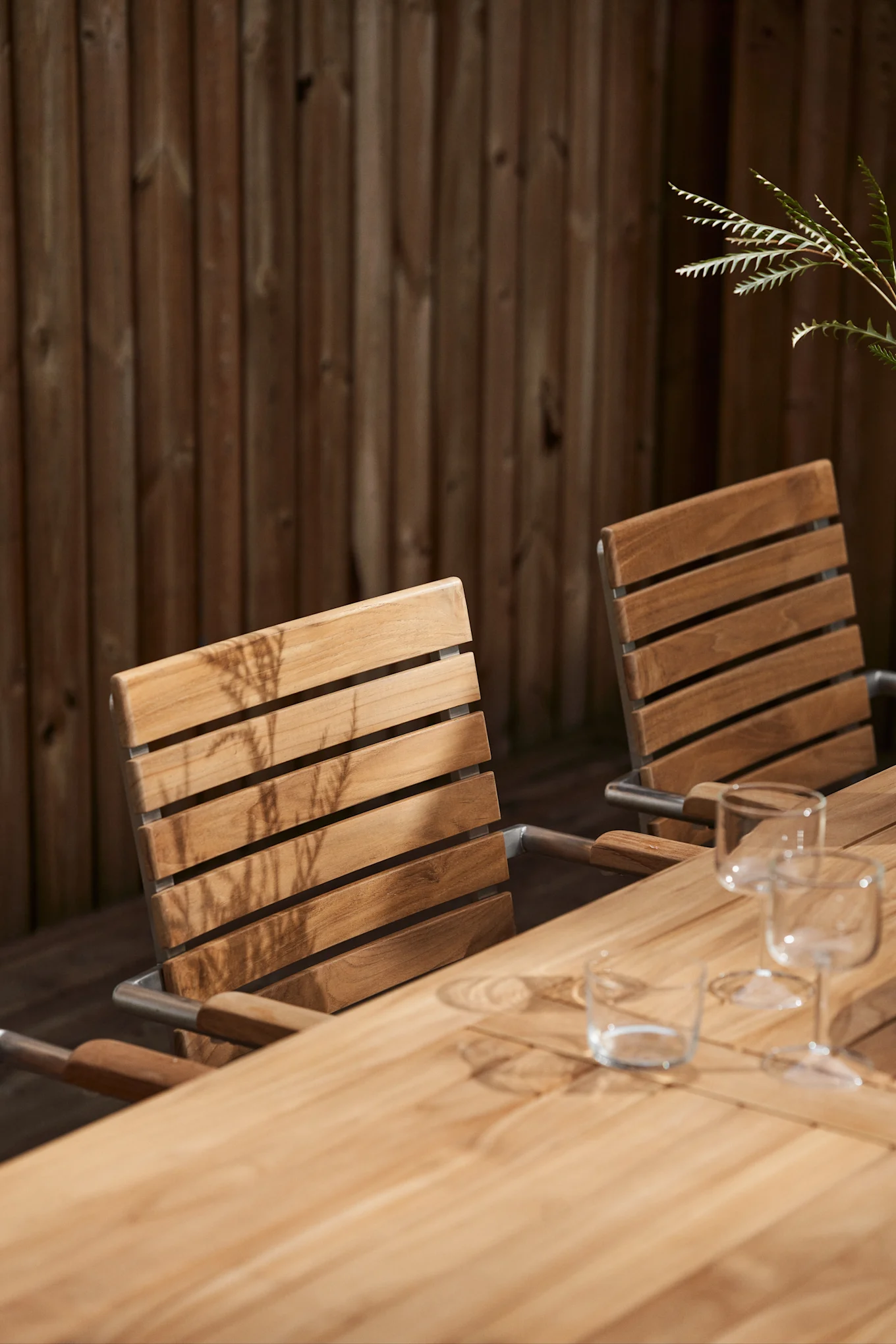Outdoor teak dining chairs and table with glasses, plant, and wooden fence in the background.