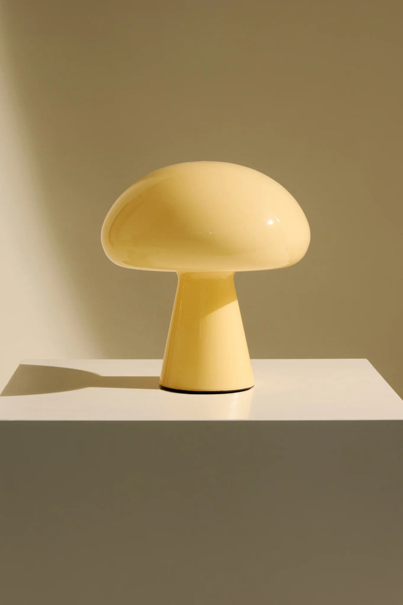 A pale yellow mushroom-shaped lamp on a white pedestal against a light brown background.