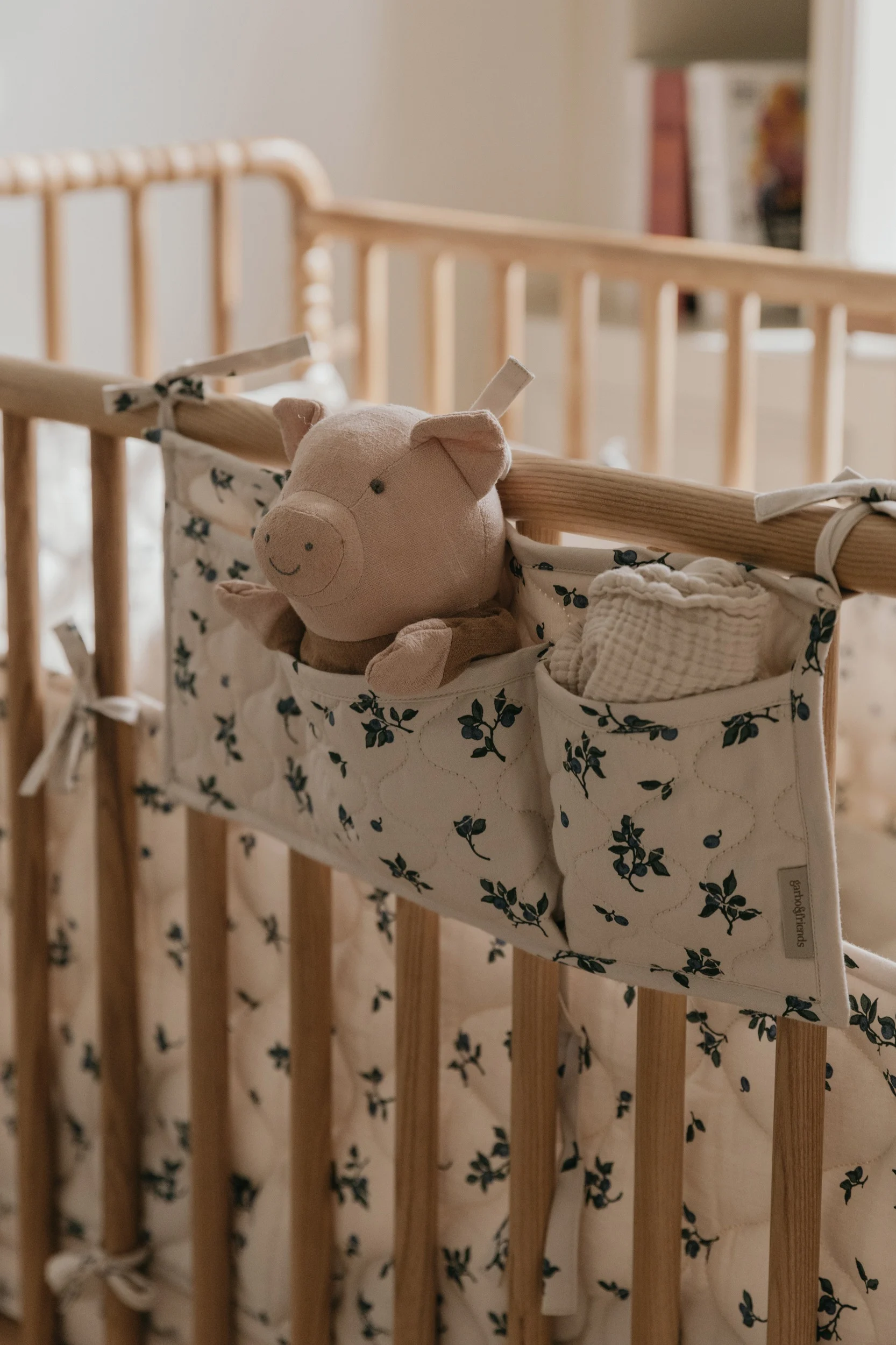 A plush pink pig toy and a white muslin cloth in a cream and blue floral organizer hanging on a wooden baby crib.