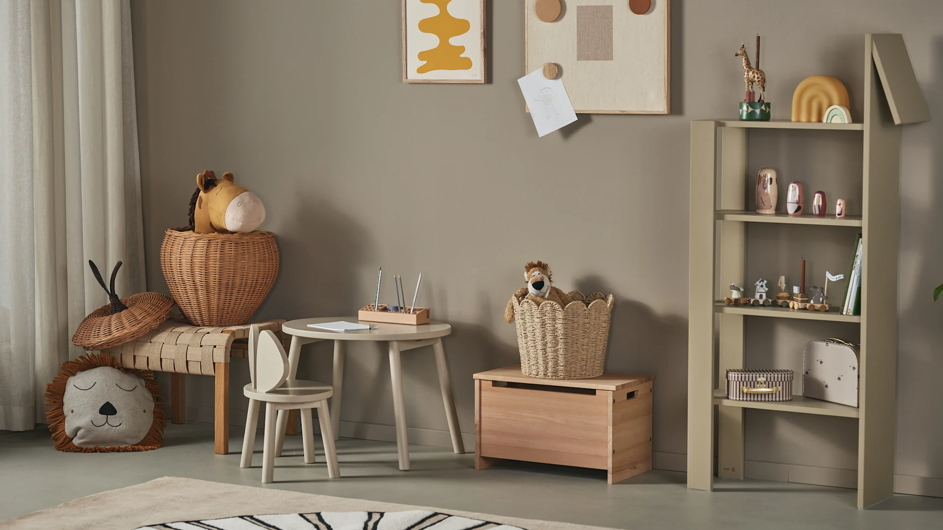 A child's room with a small wooden table, chair, wicker baskets with plush toys, lion pillow, and a bookshelf with a toy train and decor.