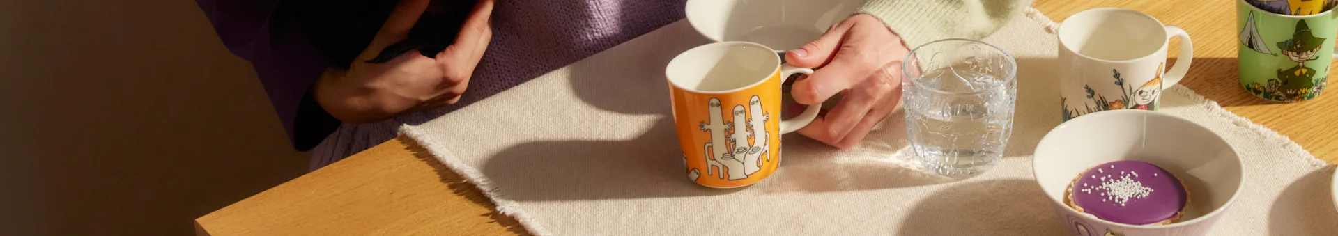Overhead shot of a person holding an orange mug, next to a glass of water, other patterned mugs, and a purple tart on a wooden table.