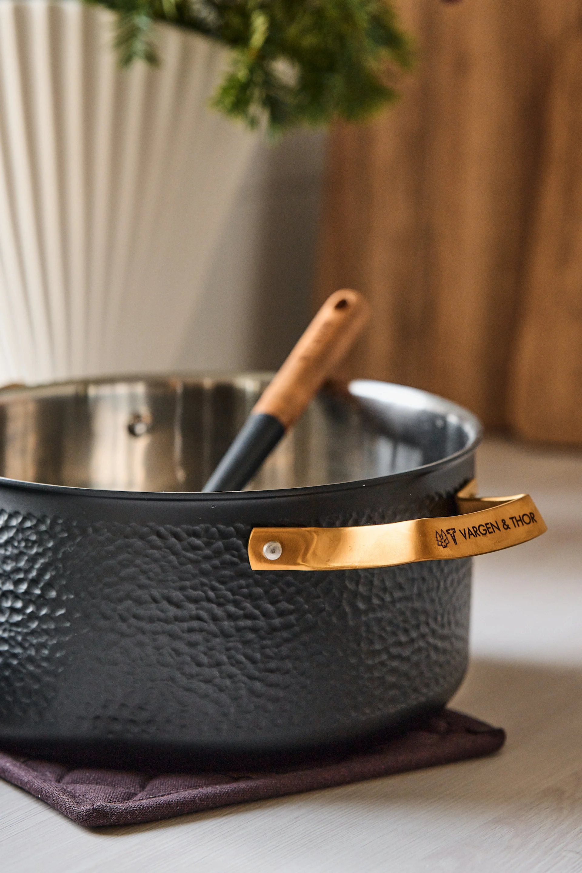 Black textured pot with gold-colored handles and a wooden-handled utensil, resting on a dark placemat on a light wooden surface.