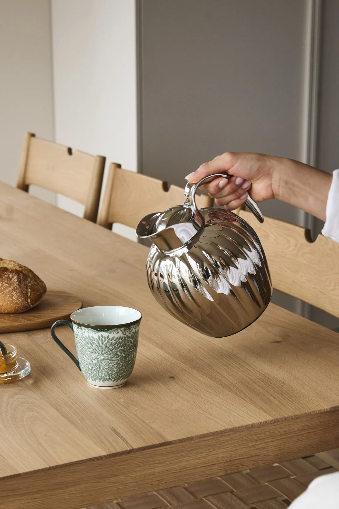 A hand pours from a silver fluted thermal carafe into a green patterned mug on a wooden table with bread and jam.