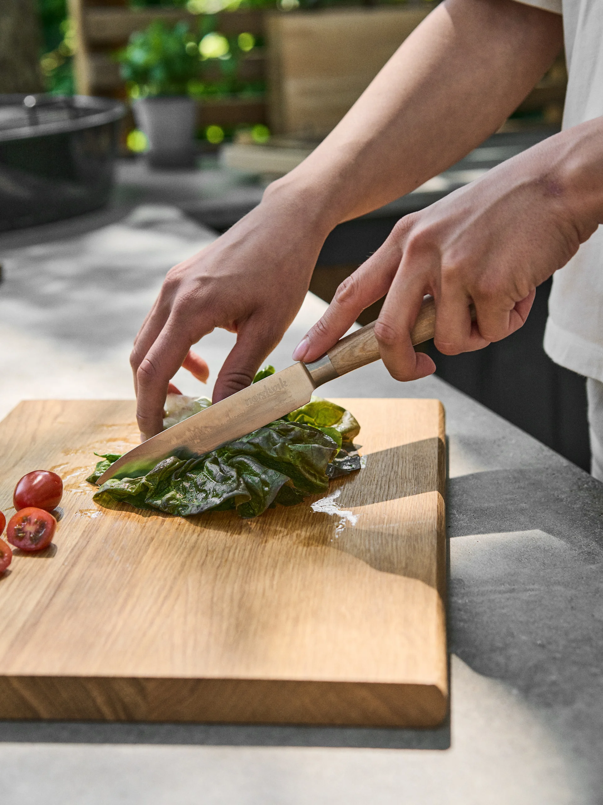 Hands cutting a green leaf with a knife on a wooden board, next to cherry tomatoes.
