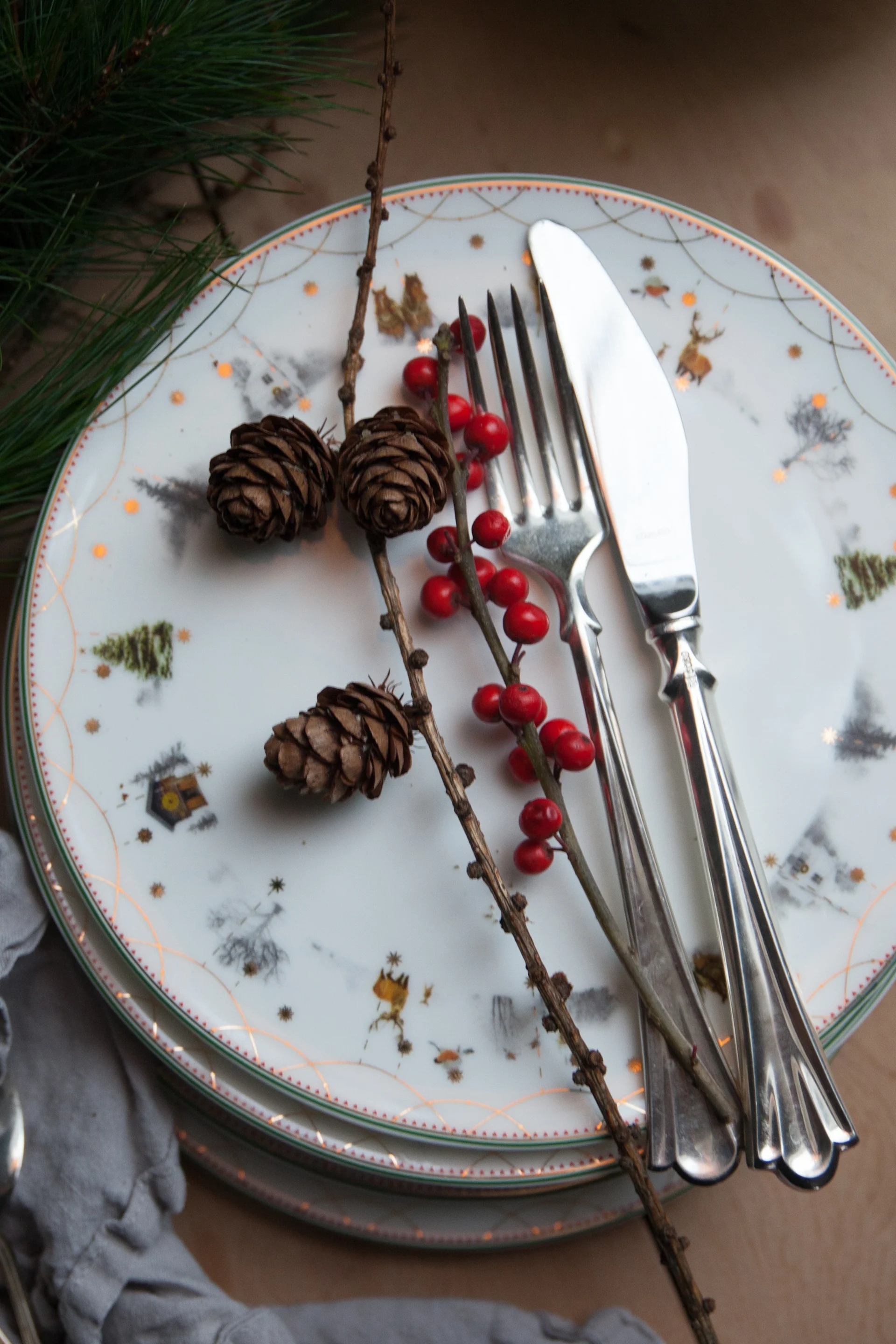 A festive Christmas table setting with stacked plates decorated with reindeer and trees, silver fork and knife, pinecones, and red berries.