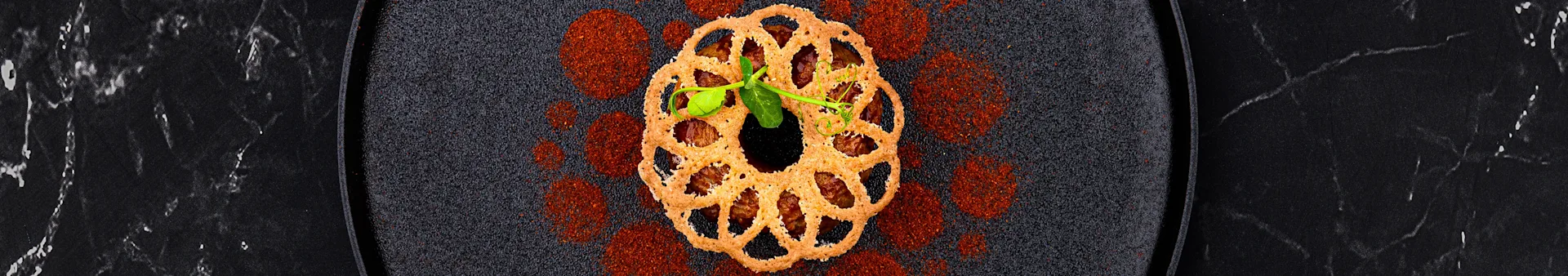 Crispy ring-shaped cracker with brown filling and green sprout on a dark, spiced plate.