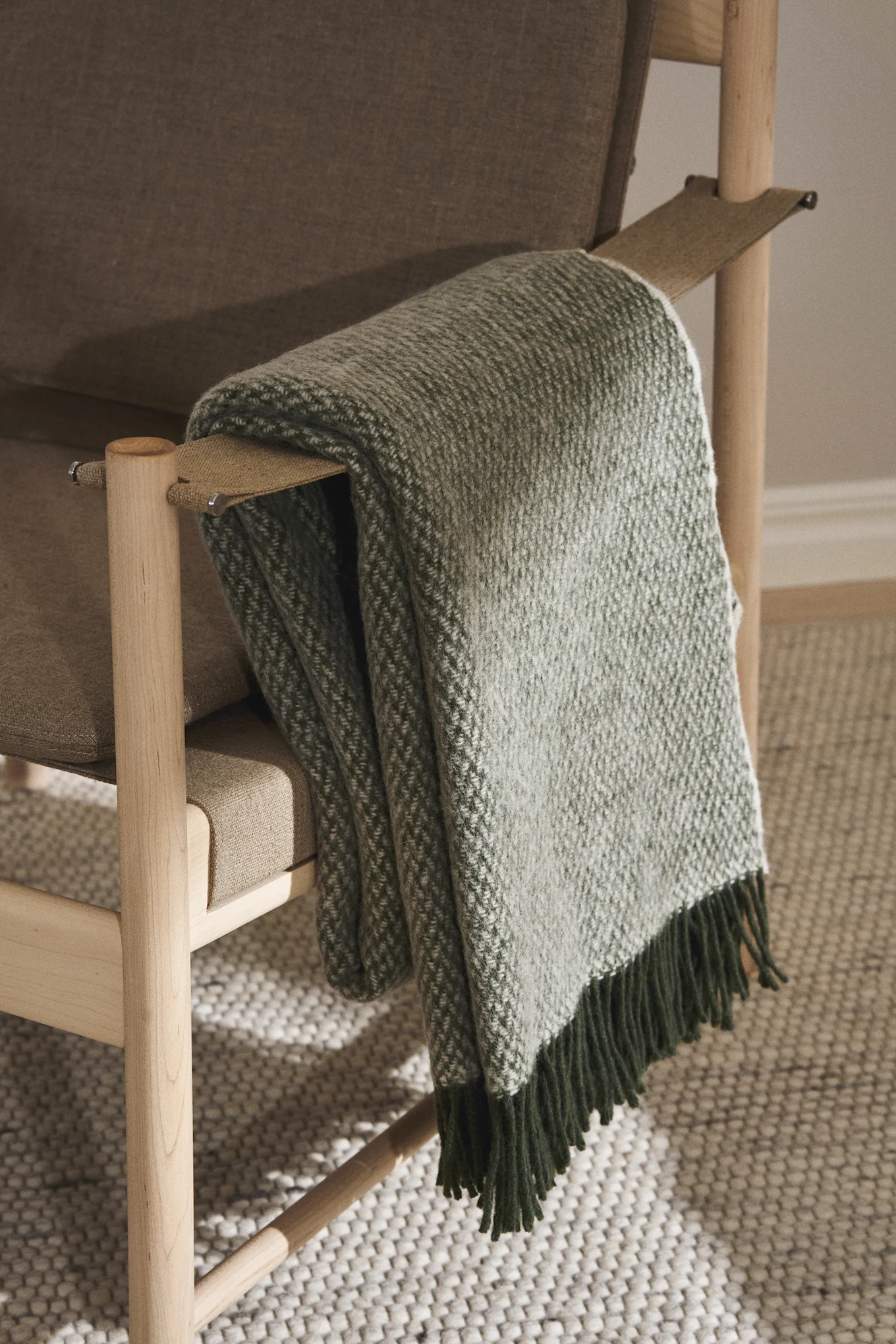 A green and white patterned blanket with dark green fringes draped over the armrest of a wooden armchair with a beige cushion.