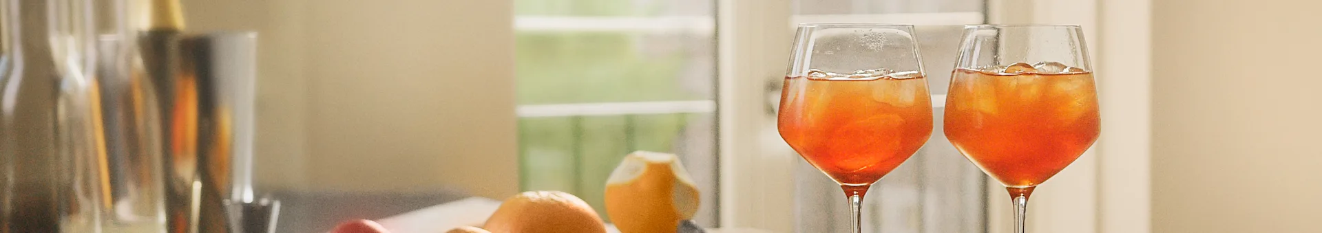 Two glasses of an orange drink with ice, with blurred fruit and a window in the background.