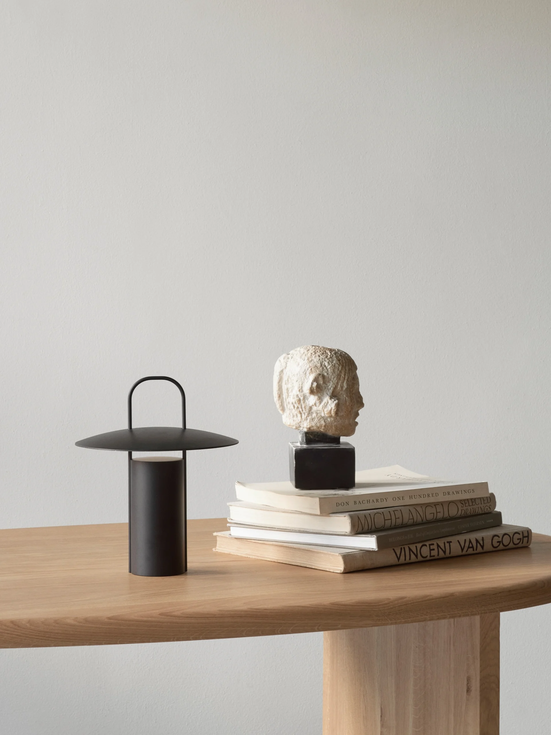 A black portable lamp and a small bust on stacked books on a light wooden table.