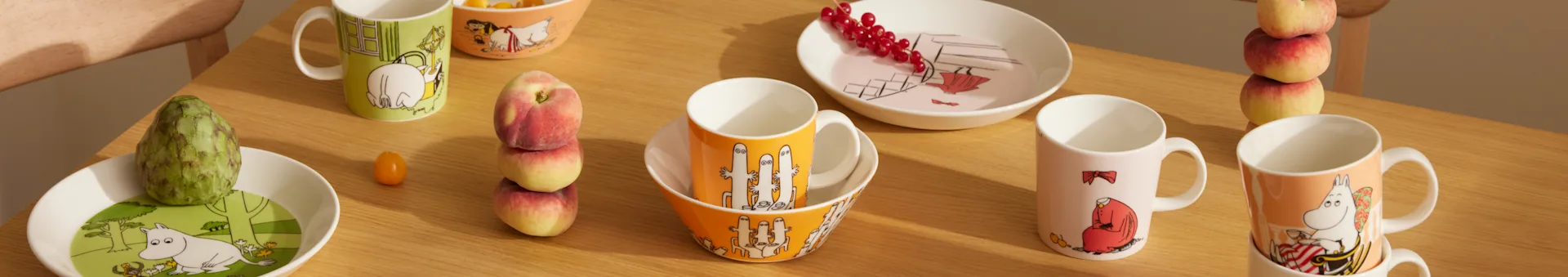 Moomin dinnerware in various colors and designs, featuring mugs, bowls, and plates, alongside fresh fruit on a wooden table.