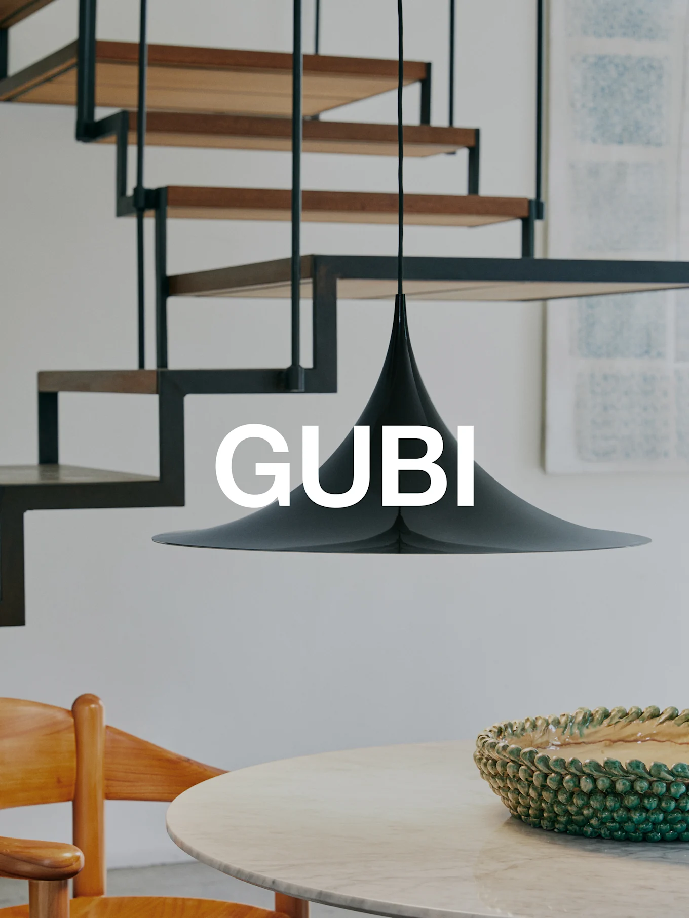 A black conical pendant lamp over a marble table with a green bowl and a wooden chair. A modern staircase is in the background. Text 'GUBI'.