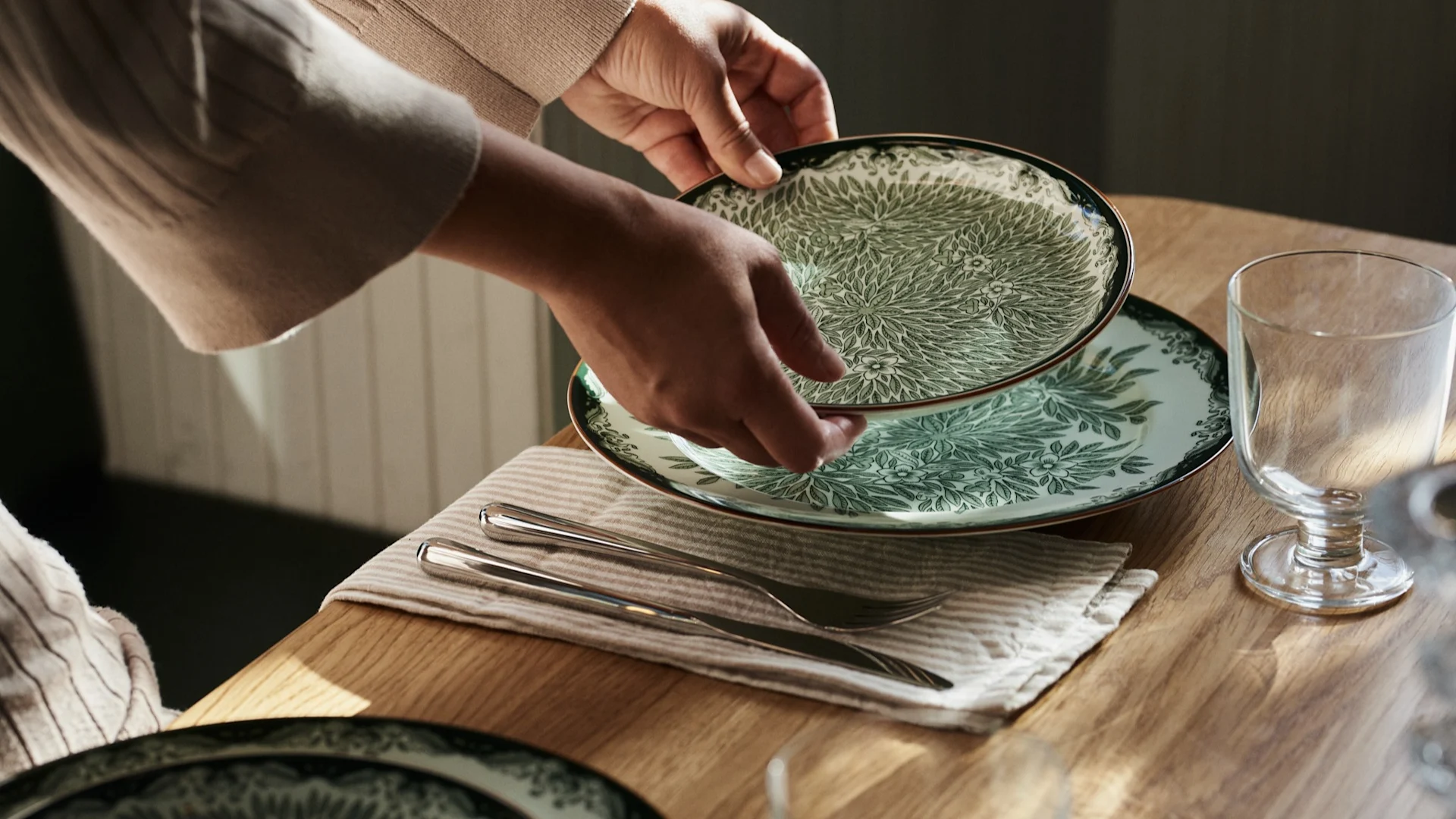 Hands placing a green and white patterned plate on a wooden table with cutlery and a glass.