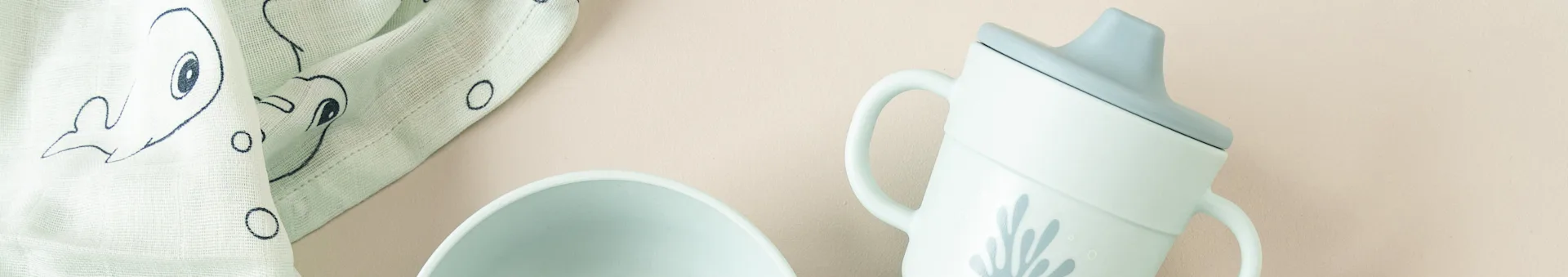 Light green baby essentials: a whale-patterned muslin cloth, a bowl, and a sippy cup with a coral design, all on a beige background.
