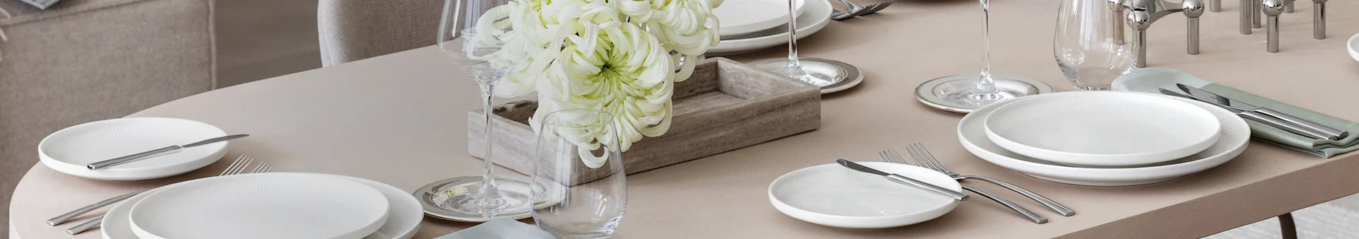 An elegantly set dining table with white plates, silver cutlery, wine and water glasses, and a centerpiece of large white flowers in a wooden box.