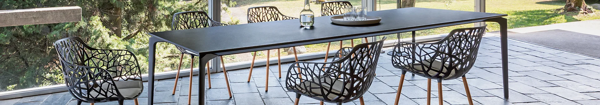 Outdoor dining set: long dark table, six chairs with tree-branch backs, light cushions, wooden legs, on a tiled patio by a garden.