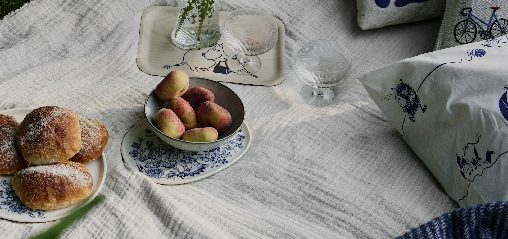 Picnic scene on a white blanket with bread rolls, peaches, drinks, and cushions featuring Moomin and bicycle patterns.
