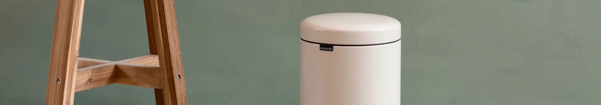 An off-white cylindrical trash can with a rounded lid and a black band, next to a light wooden stand against a green wall.