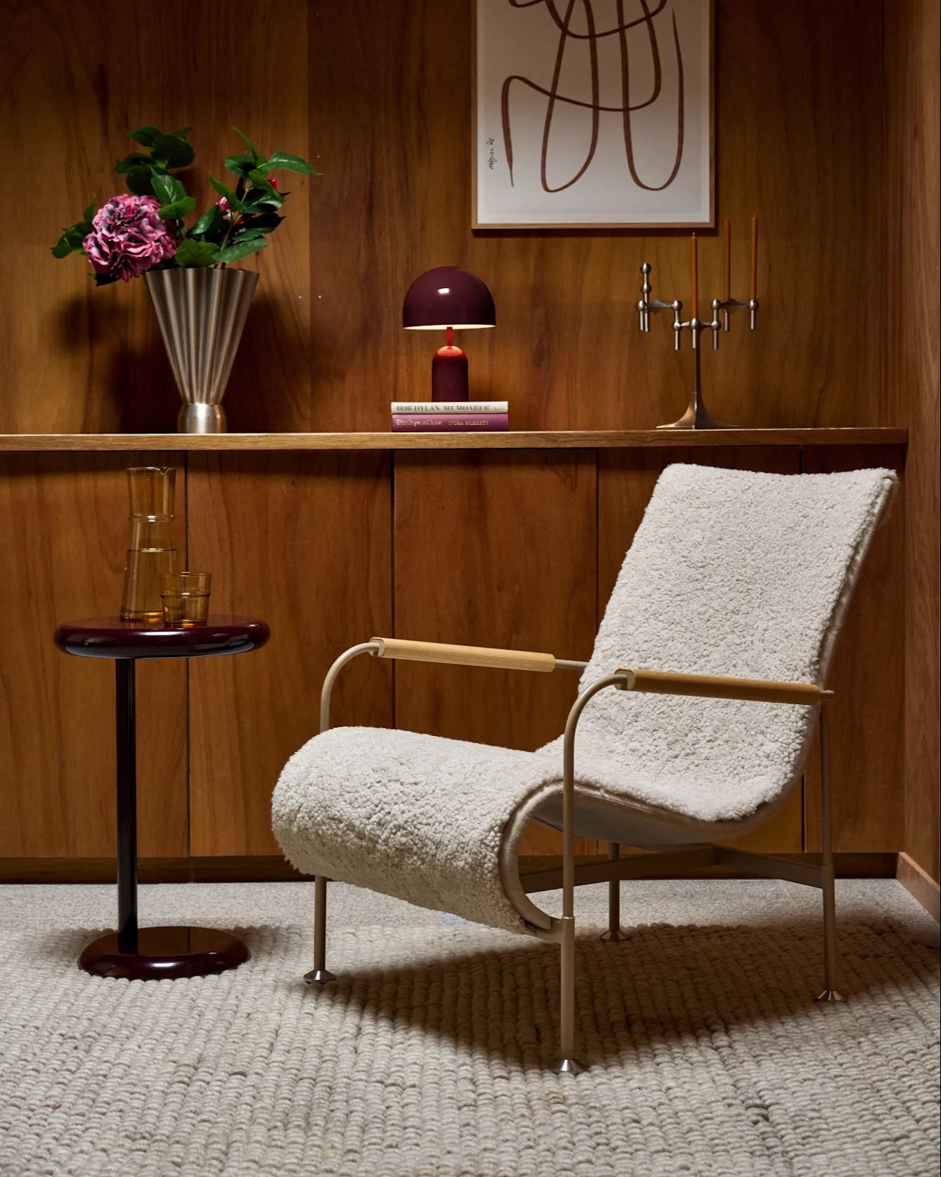 Wood-paneled room with fluffy armchair, red side table, shelf with vase, lamp, candelabra, and abstract art.