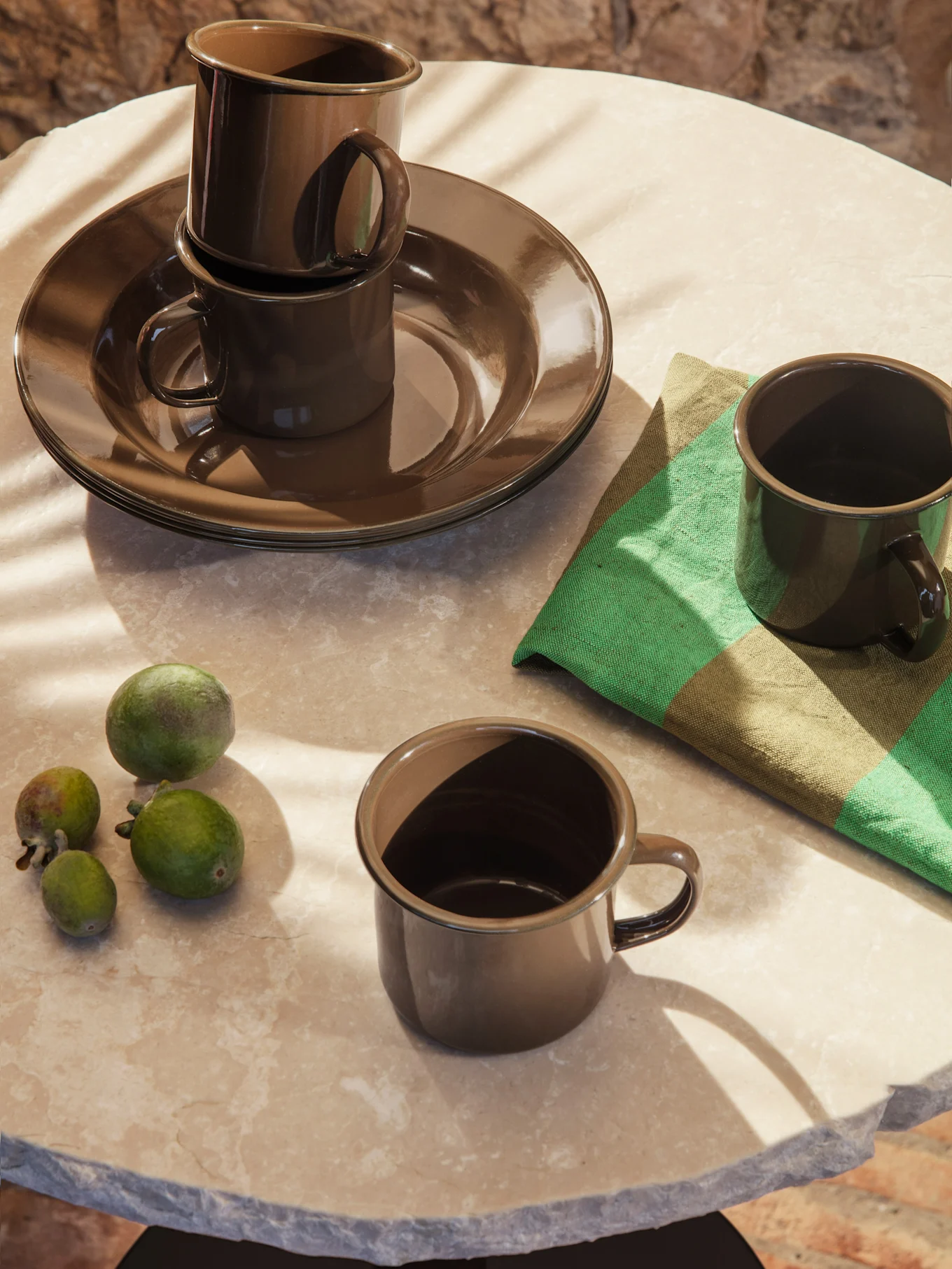 Dark brown enamelware cups and plates on a marble table with green feijoa fruits and a striped napkin, bathed in sunlight.