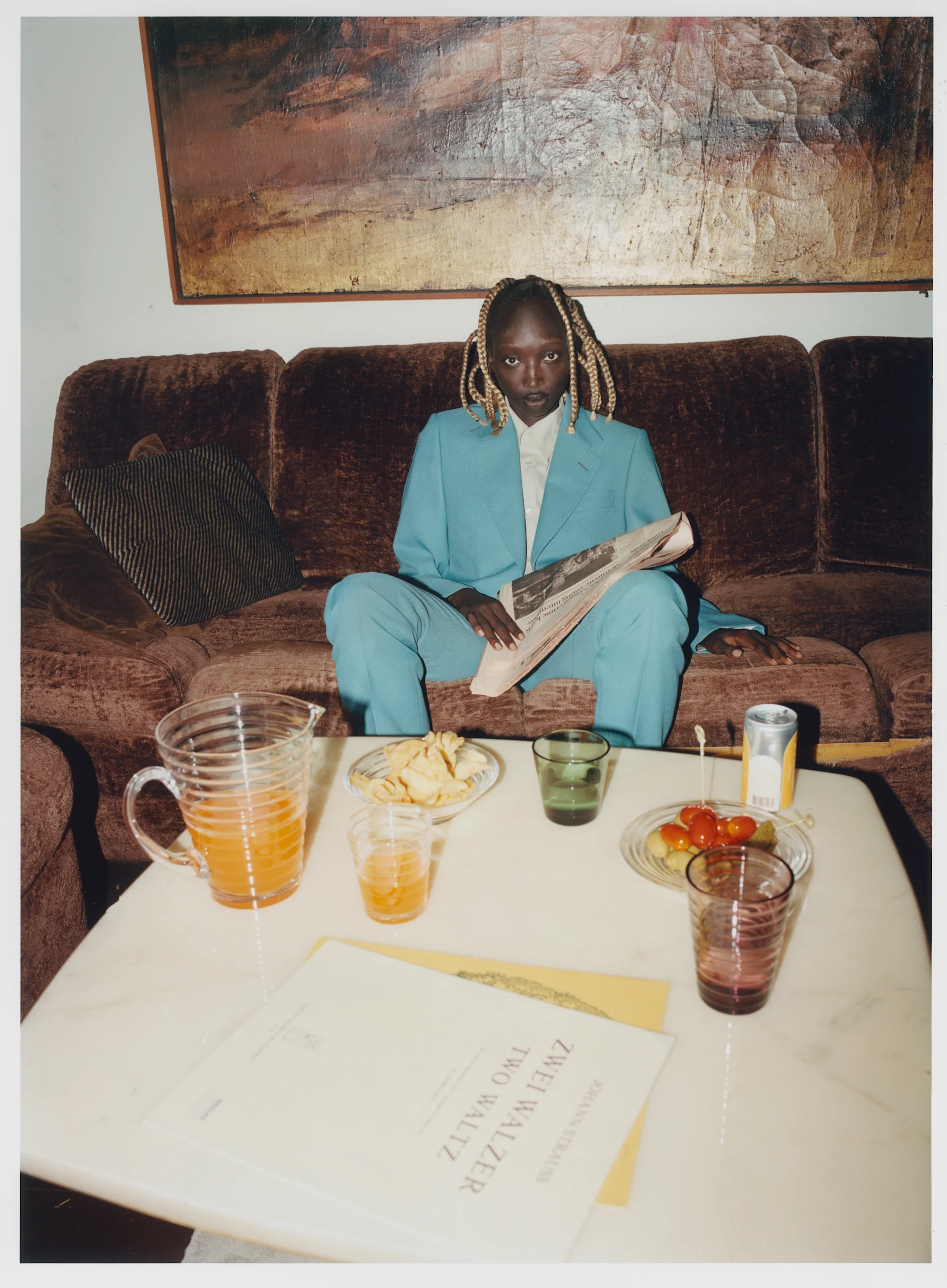 Person in a teal suit with braided hair sits on a brown couch, reading a newspaper. A coffee table holds snacks and drinks.