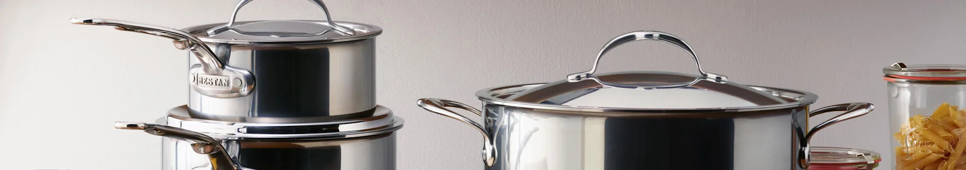 Stainless steel pots with lids, including a large one on the right and smaller, stacked ones on the left, next to a jar of pasta.