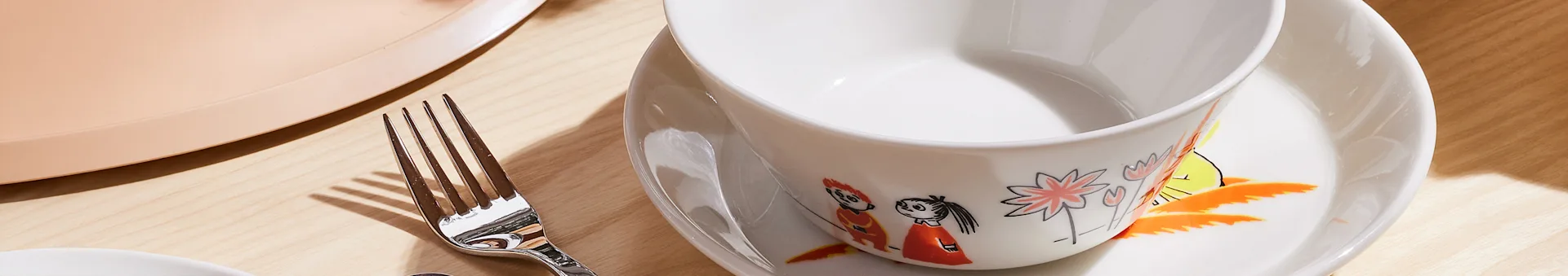 White ceramic bowl with colorful illustrations on a matching plate, next to a silver fork and a light pink tray on a wooden table.