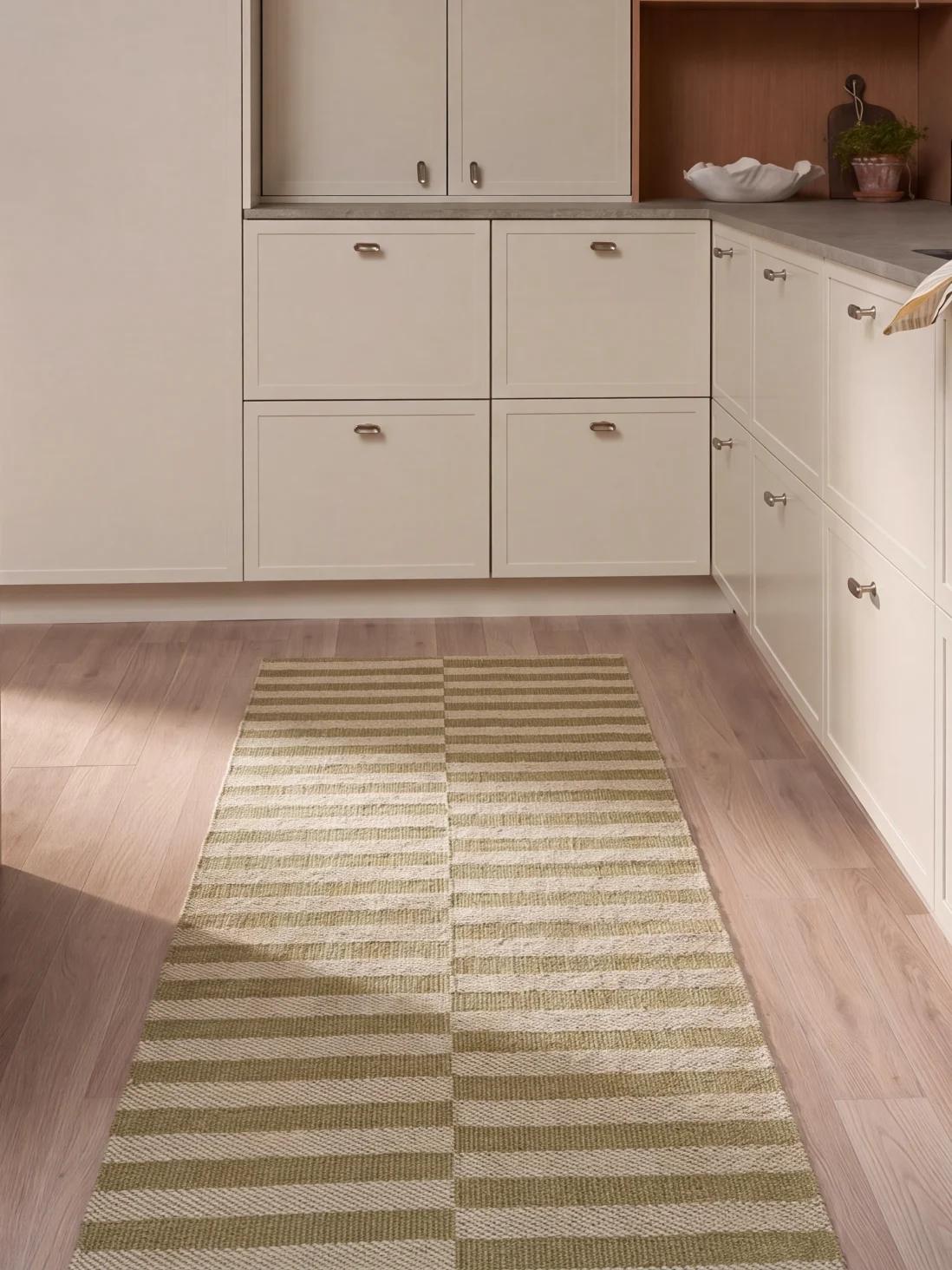 A kitchen with light cabinets, wooden flooring, and a long rectangular rug with green and white horizontal stripes.