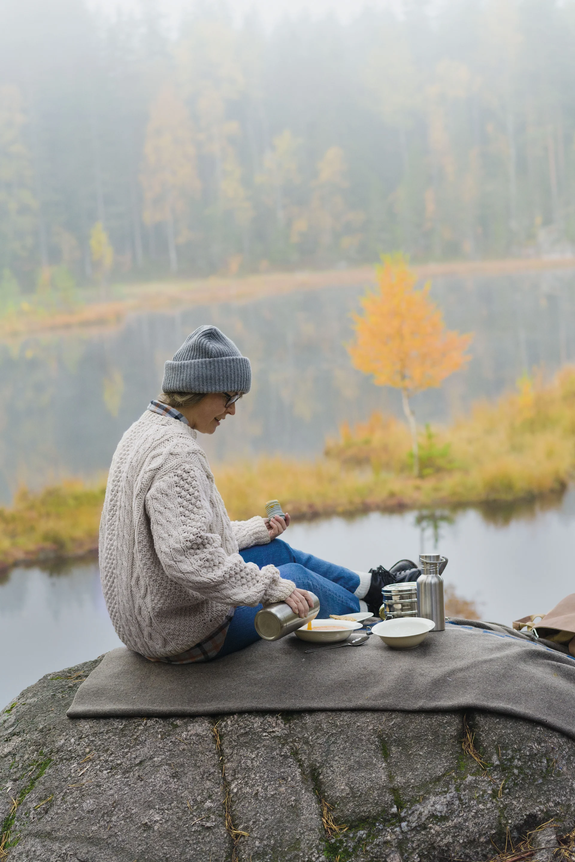 Person in a grey beanie and cream sweater pouring from a thermos into a bowl during a lakeside picnic on a foggy autumn day.