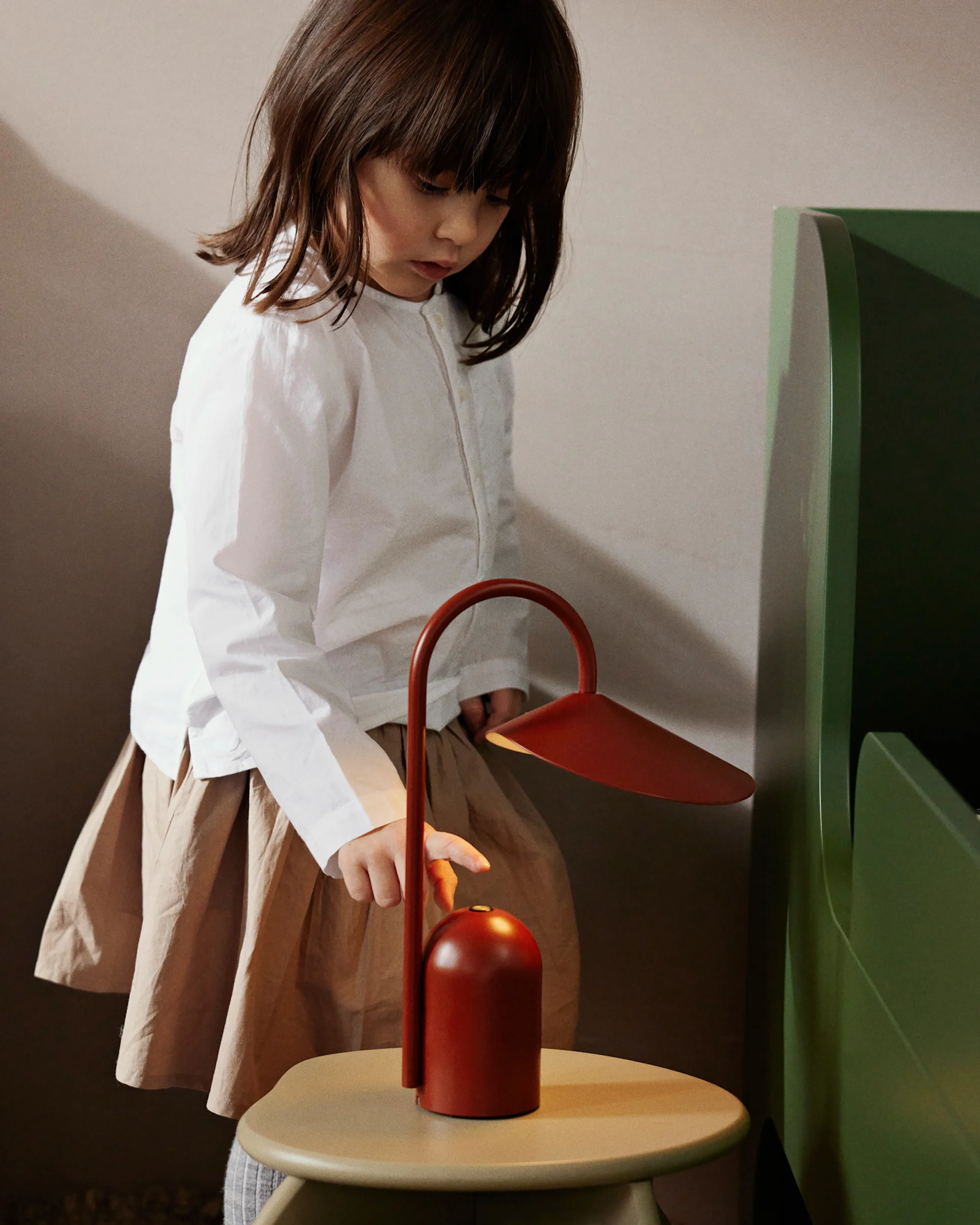A young child in a white shirt and brown skirt turns on a red portable lamp on a light-colored table.