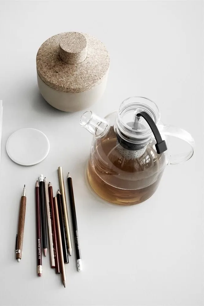 Teapot with tea, cork-lidded jar, white coaster, and pencils on a white surface.