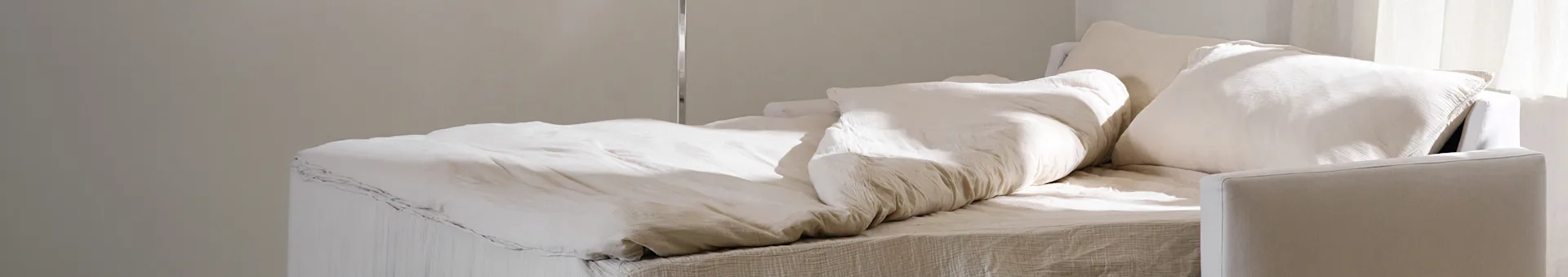 A bed with crumpled light-colored bedding and pillows, softly lit by natural light.