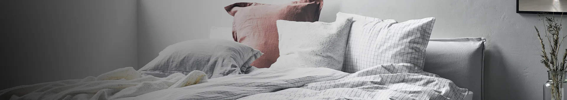 A bed with multiple pillows in shades of grey, pink, and white, with grid-patterned bedding and dried plants in a vase.