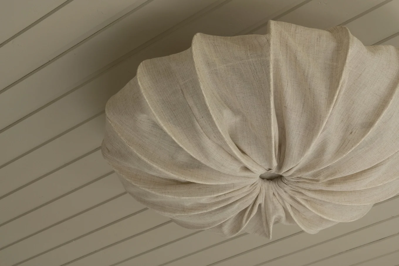A spherical, pleated light-colored fabric lampshade hanging from a light wooden slatted ceiling.
