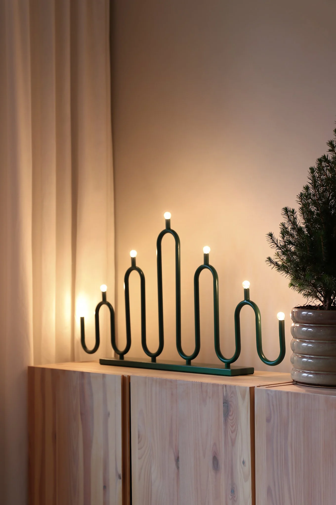 A green, wavy decorative candelabra with seven glowing bulbs sits on a light wooden cabinet next to a potted evergreen plant.