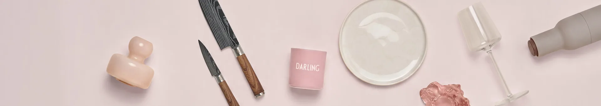 Decorative arrangement on pink: pink vase, two patterned knives, 'DARLING' candle holder, beige plate, wine glass, grey bottle with cork.
