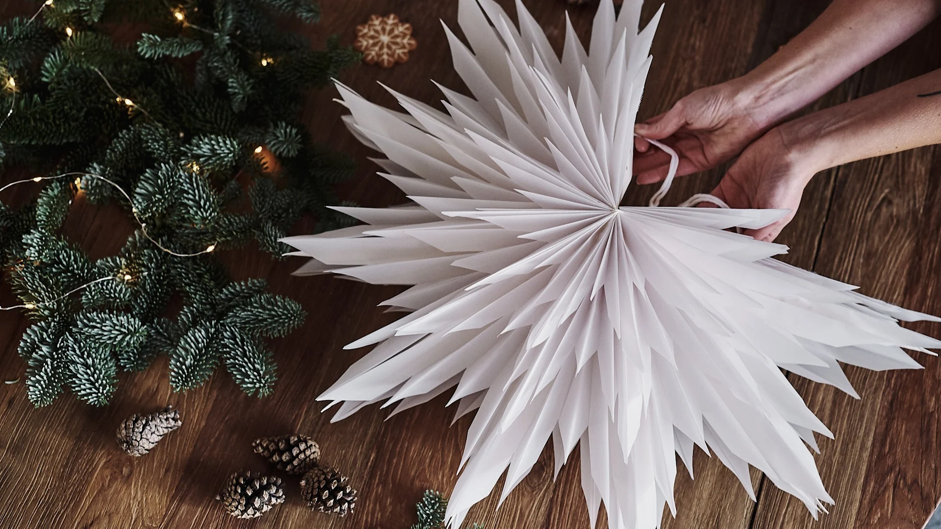 Hands assembling a white paper Christmas star, surrounded by fir branches with lights, pinecones, and a gingerbread cookie.