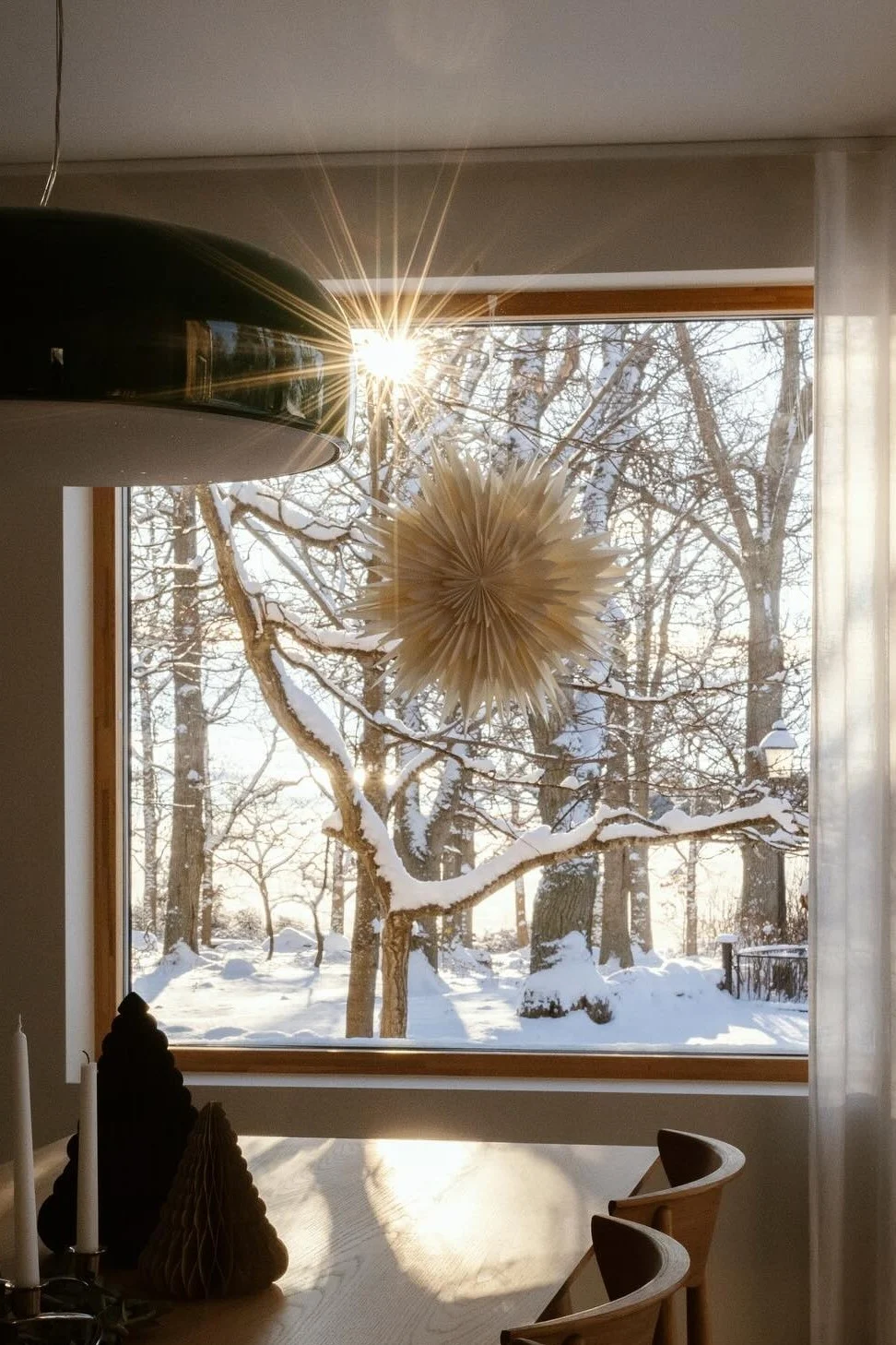 A bright winter scene through a window, with snowy trees and a sunburst. Inside, a table with candles and paper trees, and a paper star hangs in the window.