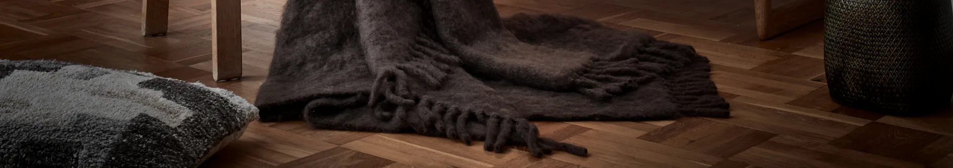Close-up of a dark brown shaggy blanket with fringes on a herringbone wooden floor, next to a patterned grey and white rug.