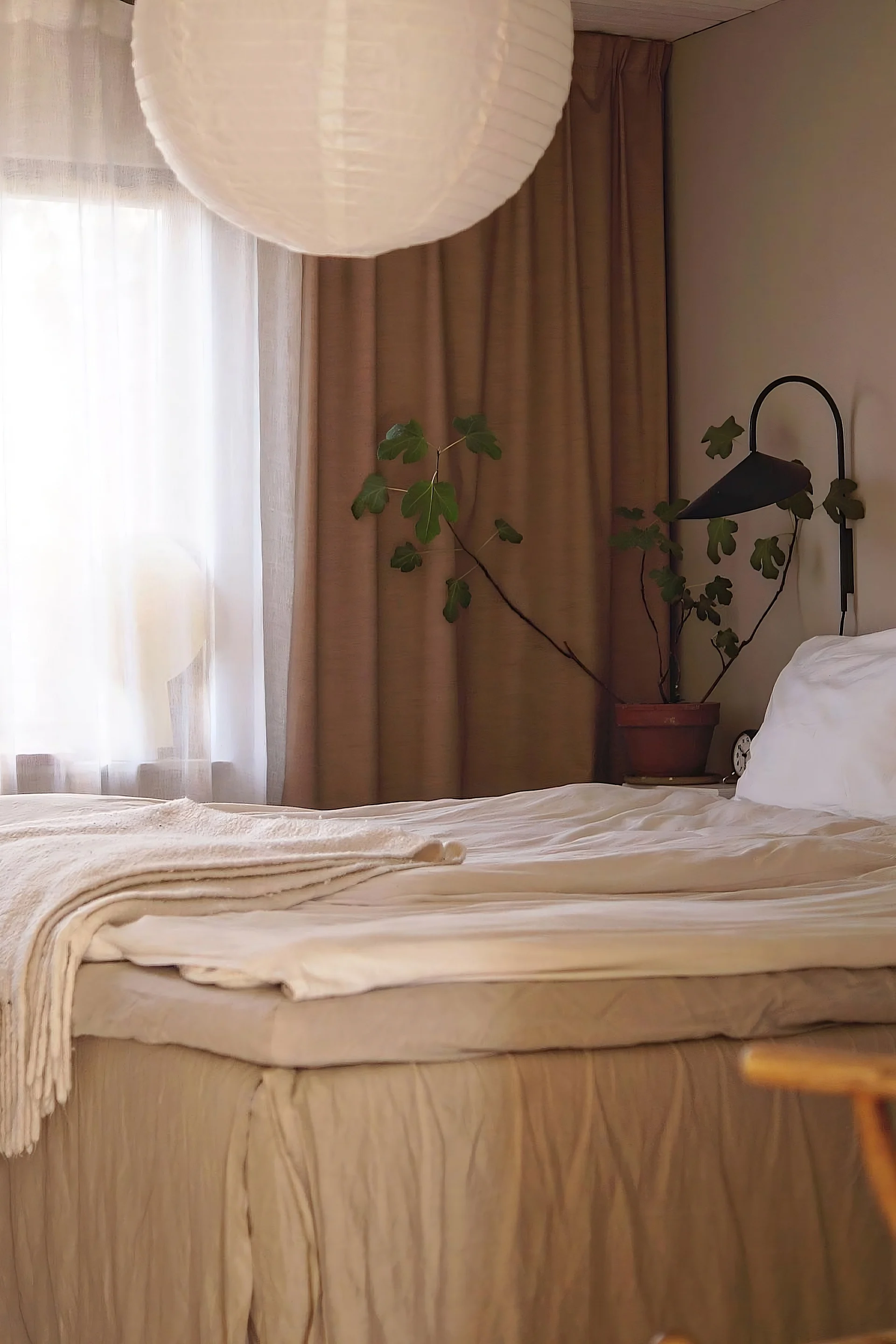 A cozy bedroom with a neatly made bed in neutral tones, a large paper lantern, a green potted plant, and a black wall lamp.