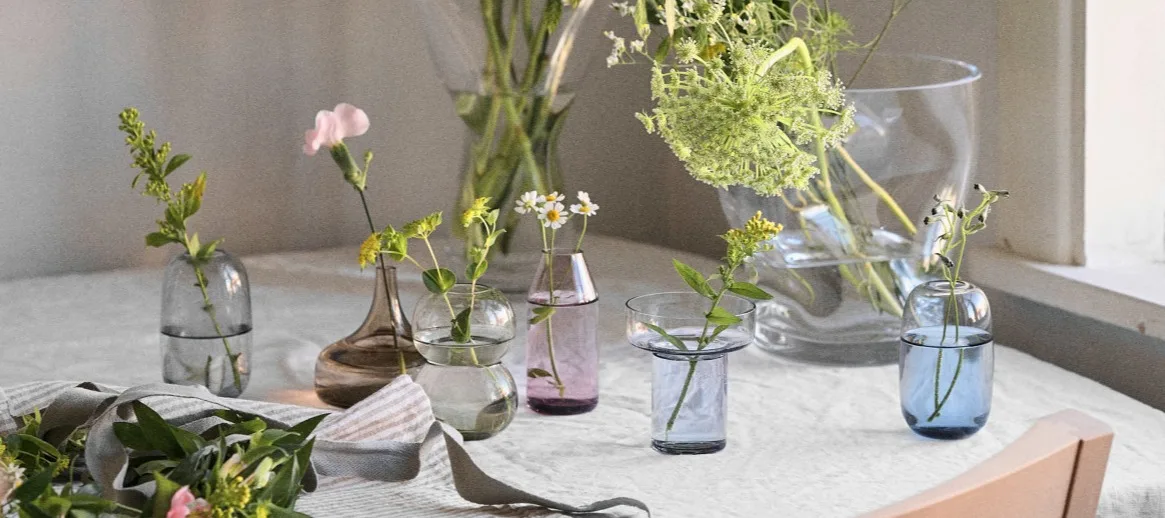 A collection of glass vases in various shapes and colors, holding different flowers, arranged on a white tablecloth.