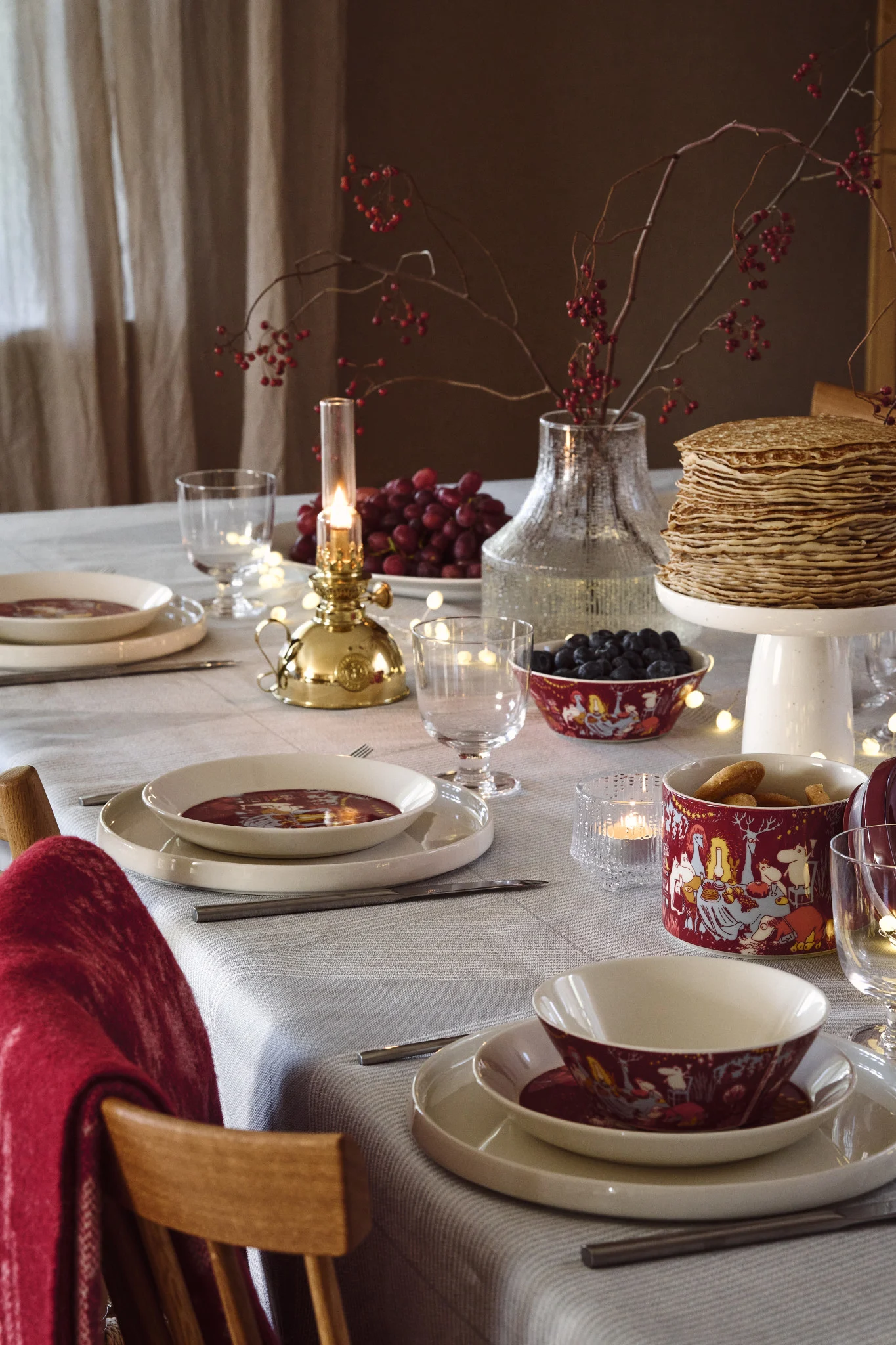 A festive dining table set with white plates, red character bowls, a stack of pancakes, fruit, and a lit oil lamp.