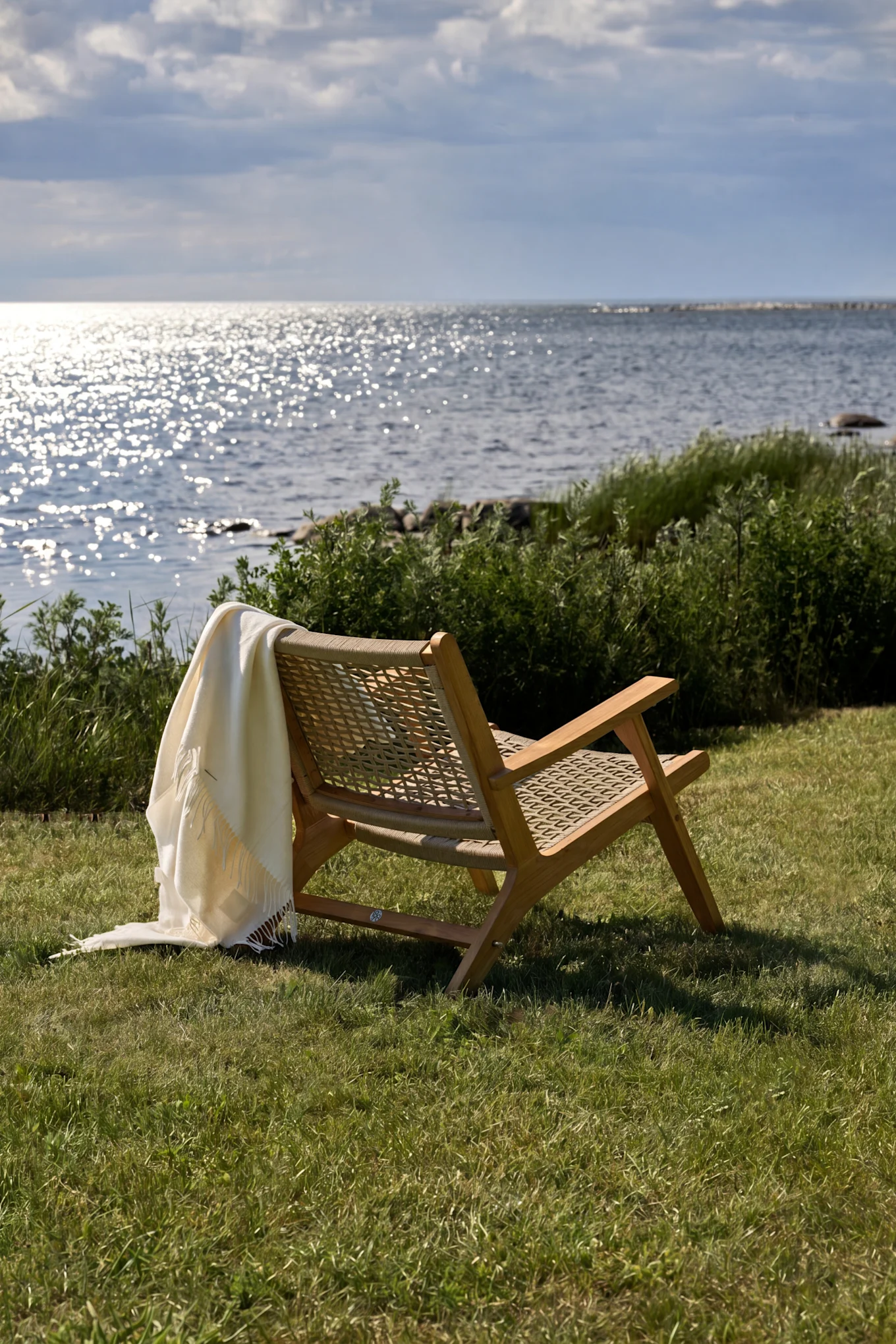 Wooden lounge chair with a white throw blanket on green grass, facing sparkling water under a cloudy sky.