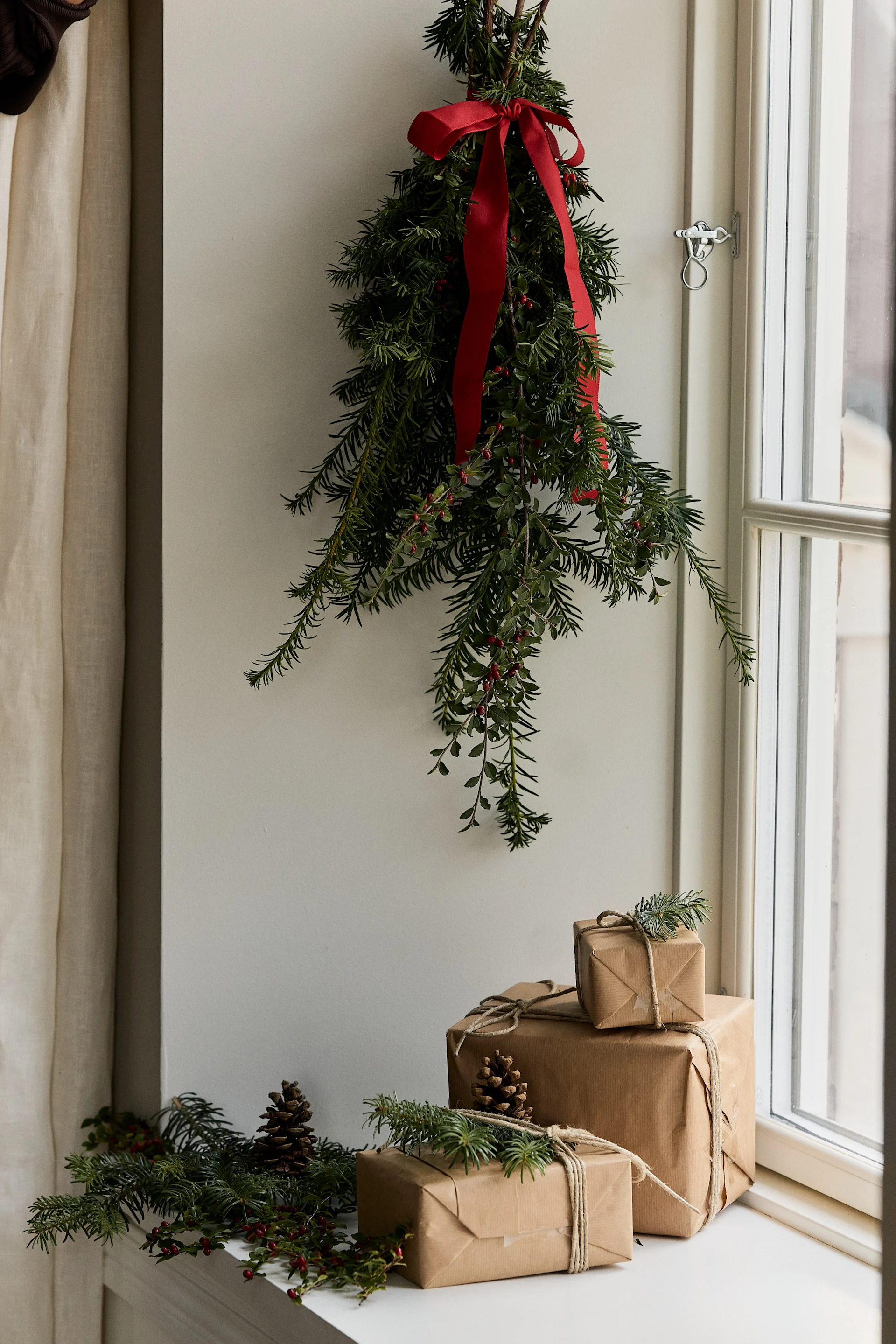 Holiday decor with hanging evergreen and red ribbon, three brown paper gifts with pinecones and sprigs on a windowsill.